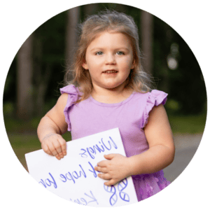 Meet Kenidee, a resilient child living joyfully despite the challenges of Dravet Syndrome. A young girl with blonde hair and a lavender dress stands outdoors on a tree-lined path, holding a white sign with handwritten text.