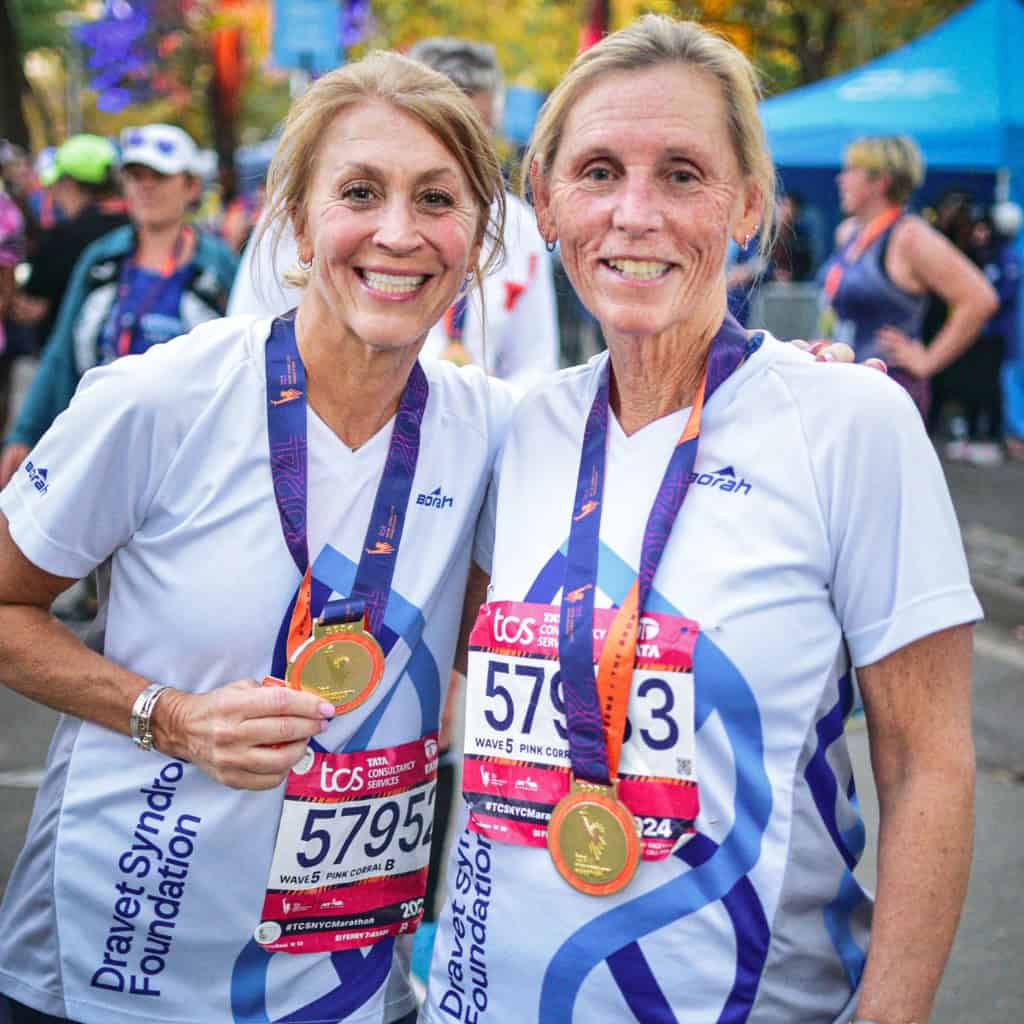 TCS New York City Marathon 3 Two women wearing purple and white marathon shirts, numbered 5795 and 5734, beam with pride as they pose with their medals. Amid a colorful sea of participants and a blue tent in the background, they celebrate completing the NYC Marathon supporting the Dravet Syndrome Foundation.